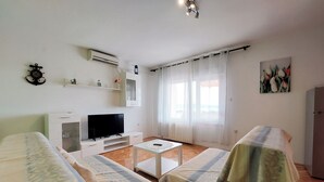 Ein modernes, helles Zimmer mit einem Flachbildfernseher, zwei Betten mit gemusterten Tagesdecken, einem kleinen Tisch und einem Fenster mit durchsichtigen Vorhängen.