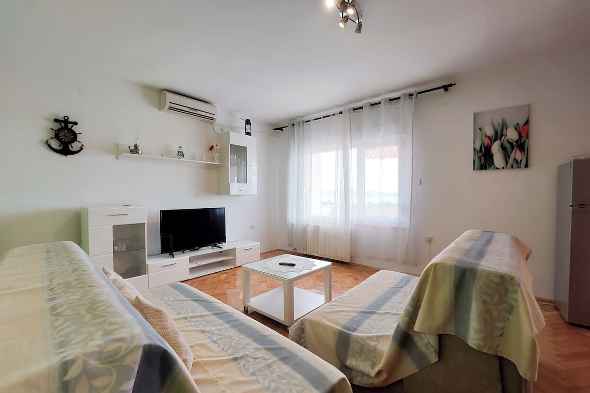 A modern, well-lit room with a flat-screen TV, two beds with patterned bedspreads, a small table, and a window with sheer curtains.