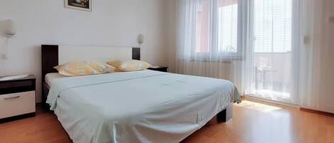 A bedroom with a bed, a nightstand, a lamp, a window with sheer curtains, and a wall-mounted picture.