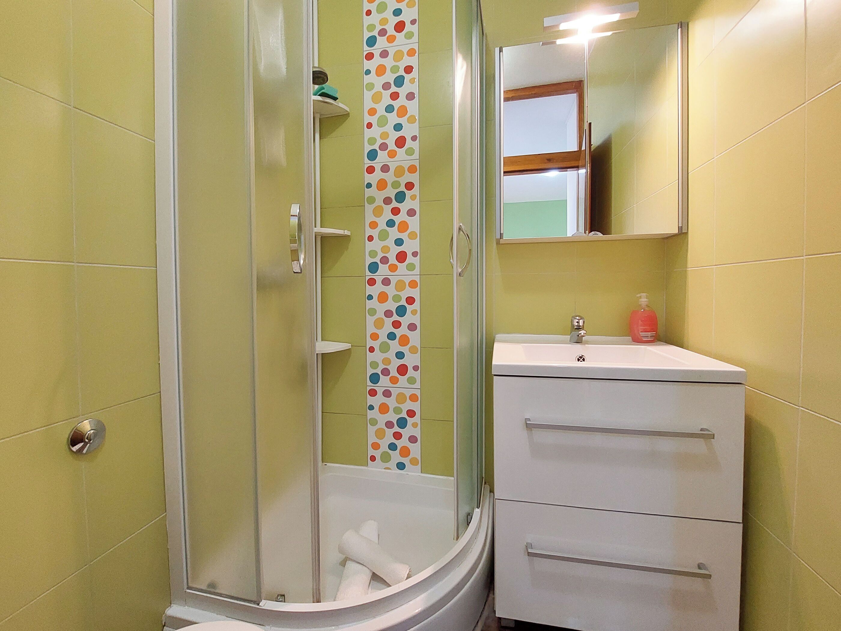 A bathroom with a glass shower enclosure, a white sink with a mirror above it, and a tiled wall with a polka dot pattern.