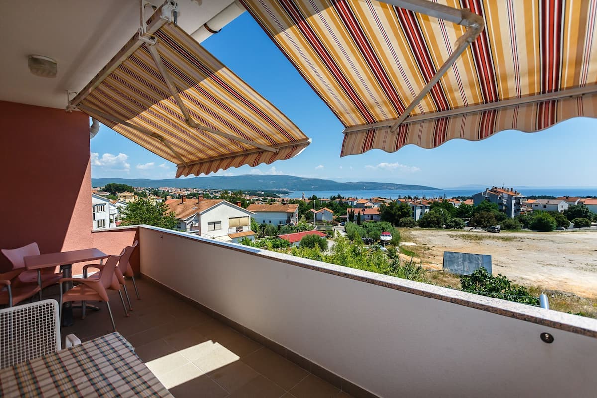 A balcony with striped awnings, a view of a coastal town, and a table with chairs.