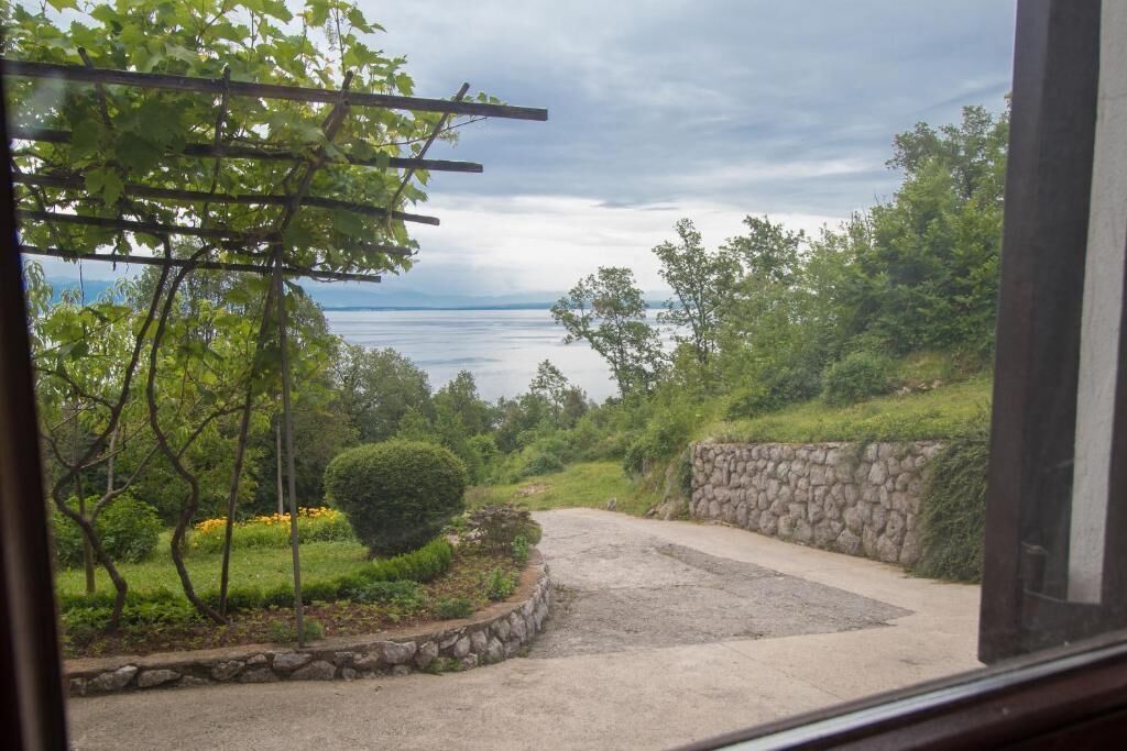 A view from a window overlooking a paved pathway leading to a body of water, surrounded by greenery and stone walls.