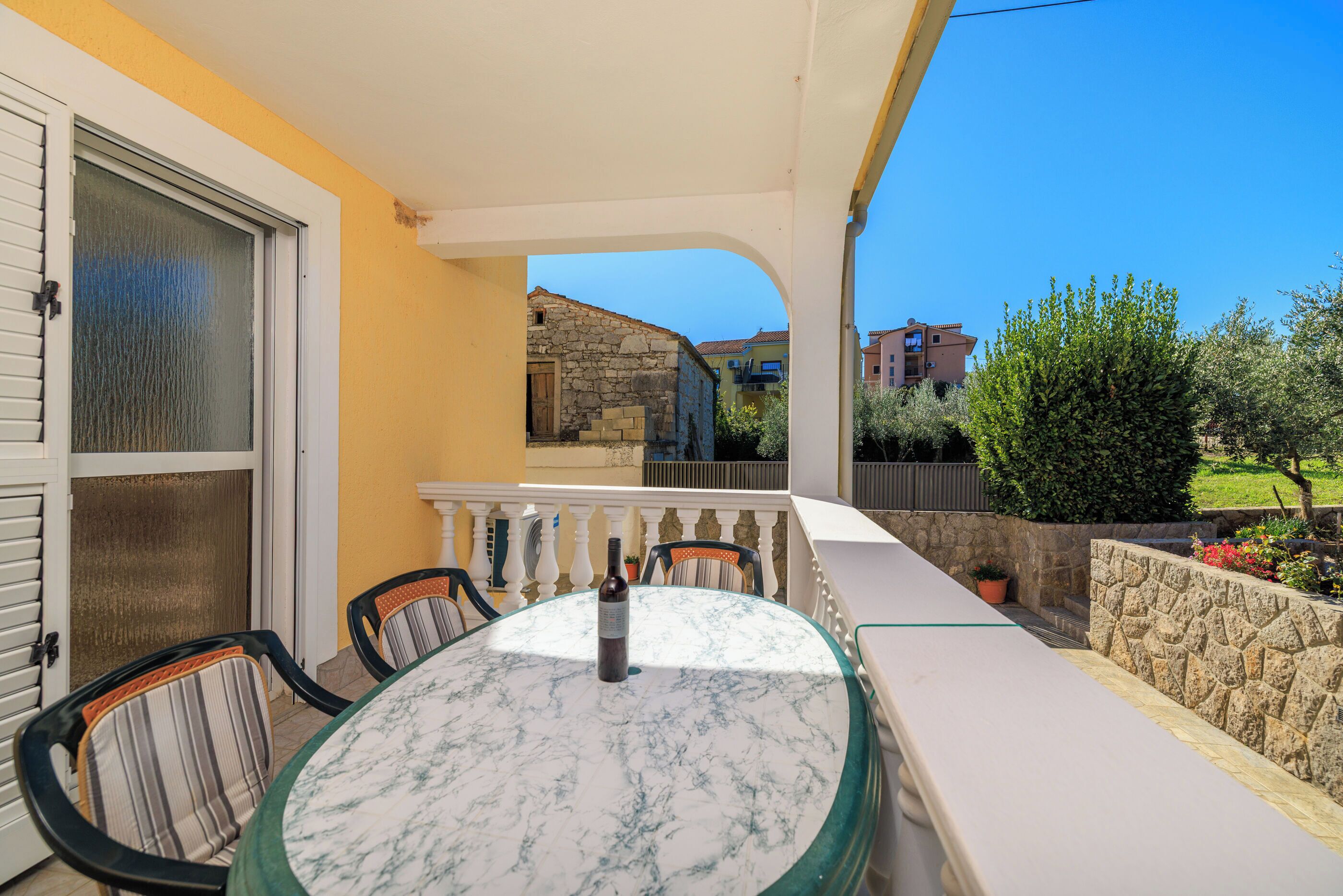 Un balcon avec une table ronde et des chaises, une bouteille posée sur la table, et une vue sur des bâtiments et de la verdure en arrière-plan.