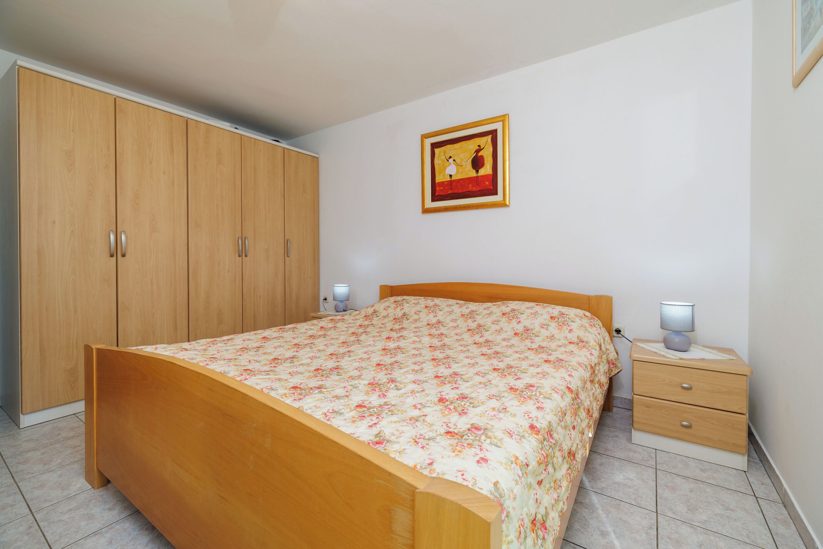 A bedroom with a wooden bed, a nightstand with two lamps, a wardrobe, and a framed picture on the wall.