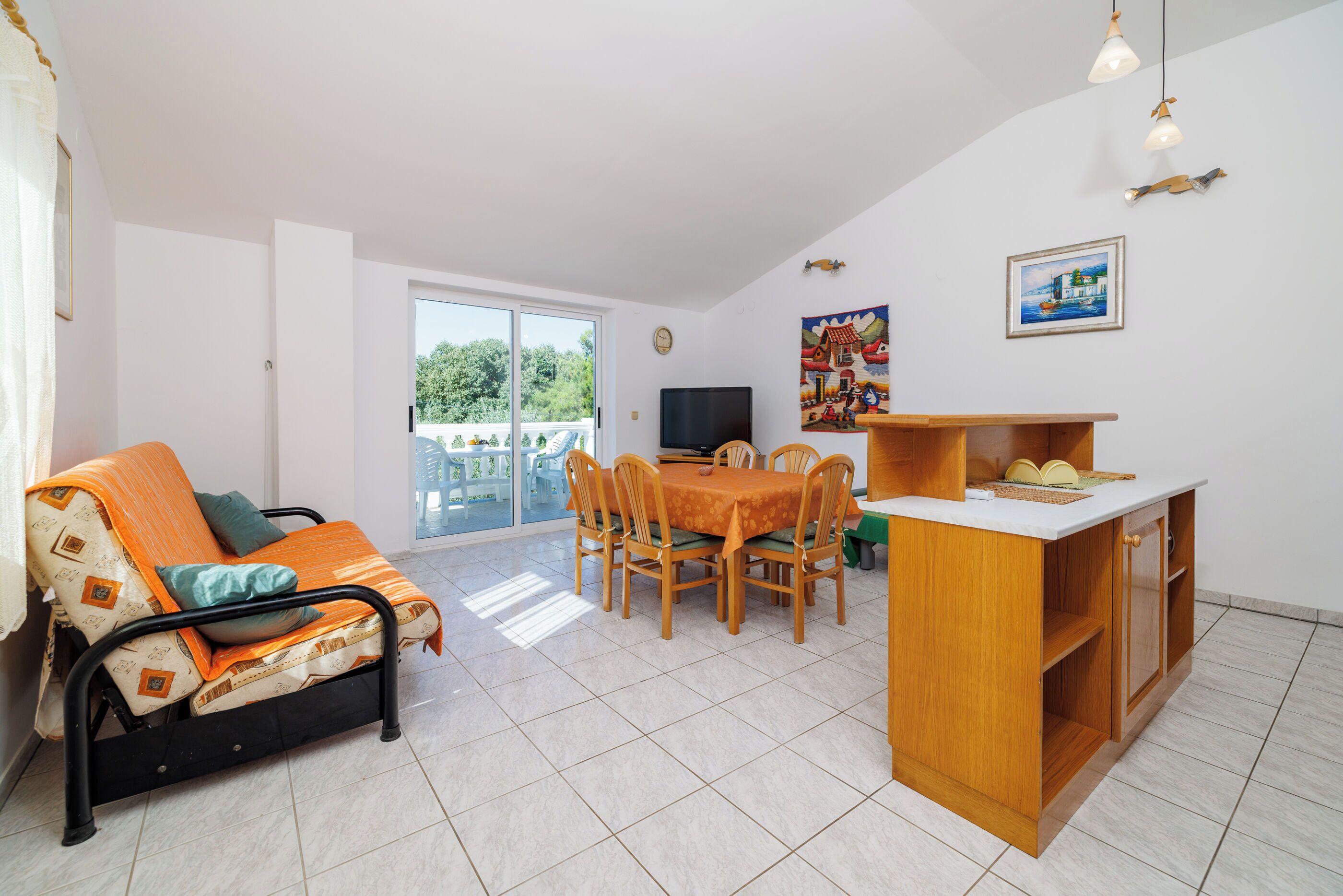 A compact living space with a sofa, dining table, chairs, a small kitchen area, and a balcony with a view of greenery.