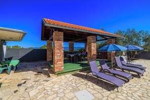 A covered outdoor seating area with stone flooring, brick pillars, and lounge chairs under umbrellas. - House Marija (Poljica)