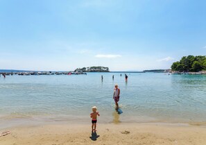Ein Strand mit klarem Wasser, einer entfernten Insel und badenden Personen.