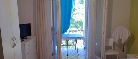 A bedroom with a bed, a television on a dresser, a balcony with a chair, and a window with curtains.