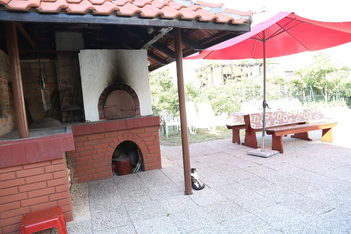 A brick outdoor kitchen with a pizza oven, a red umbrella, and a bench.