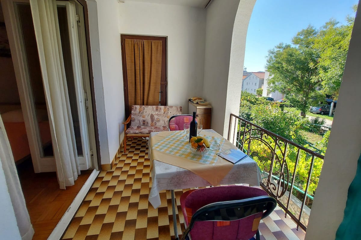 A balcony with a checkered floor, a table with a tablecloth, a chair, a sofa, and a view of greenery and buildings.