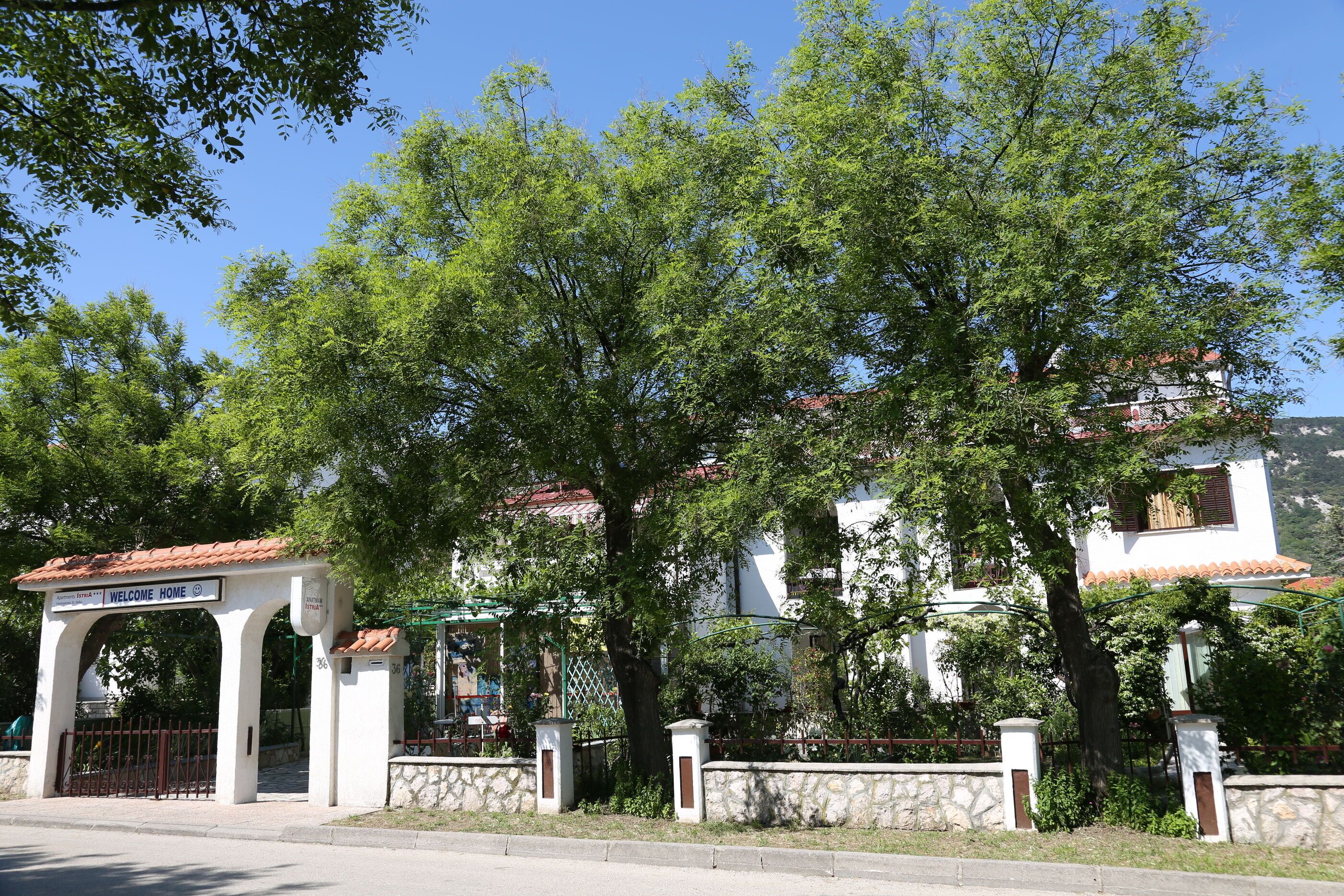A white building with a red-tiled roof, a sign reading "WELCOME HOME", and a stone wall with a gate.