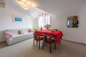 A dining area with a red tablecloth, wooden chairs, a white sofa with pink cushions, a framed picture on the wall, and a ceiling light fixture.