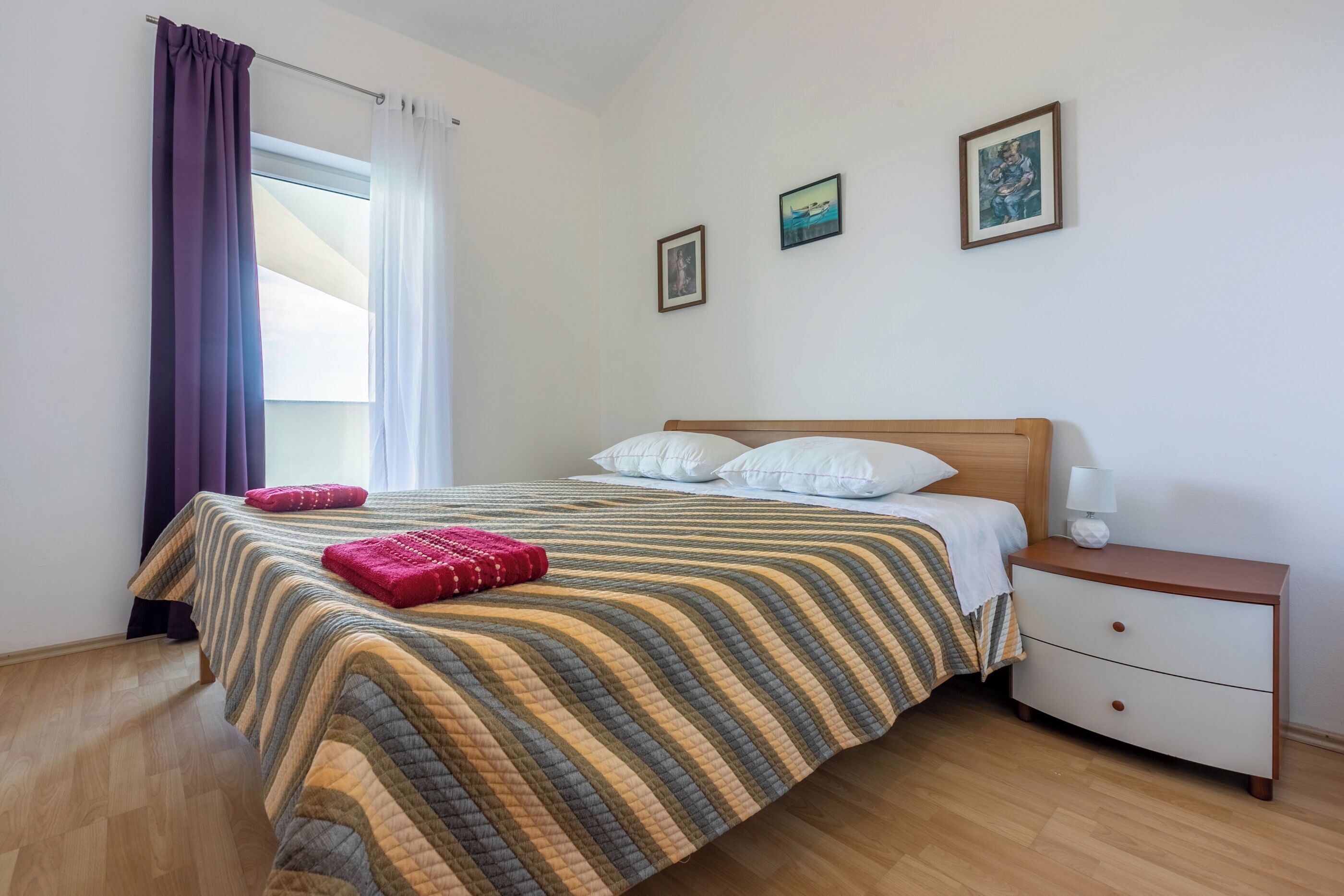 A neatly made bed with a striped bedspread, a wooden nightstand with a lamp, and a window with curtains.