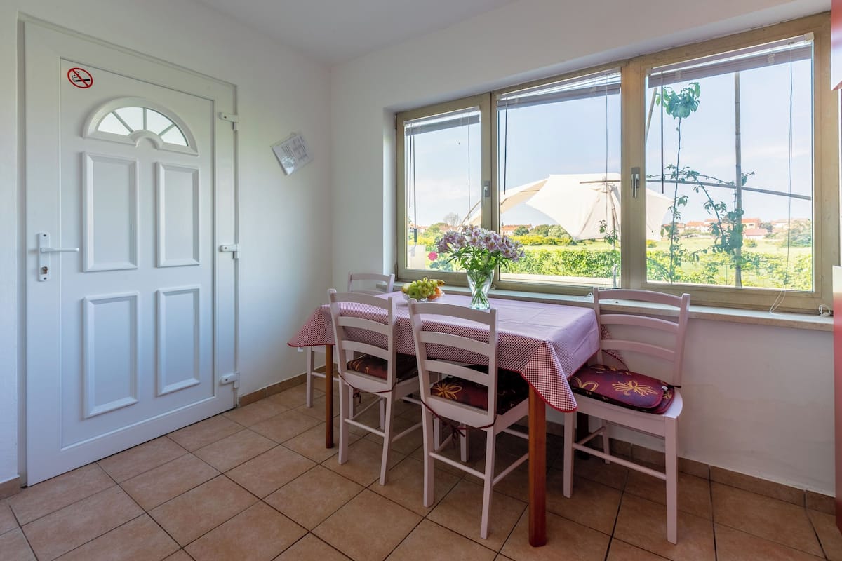 A dining area with a table set for two, a window with a view of greenery, and a white door with a no-smoking sign.