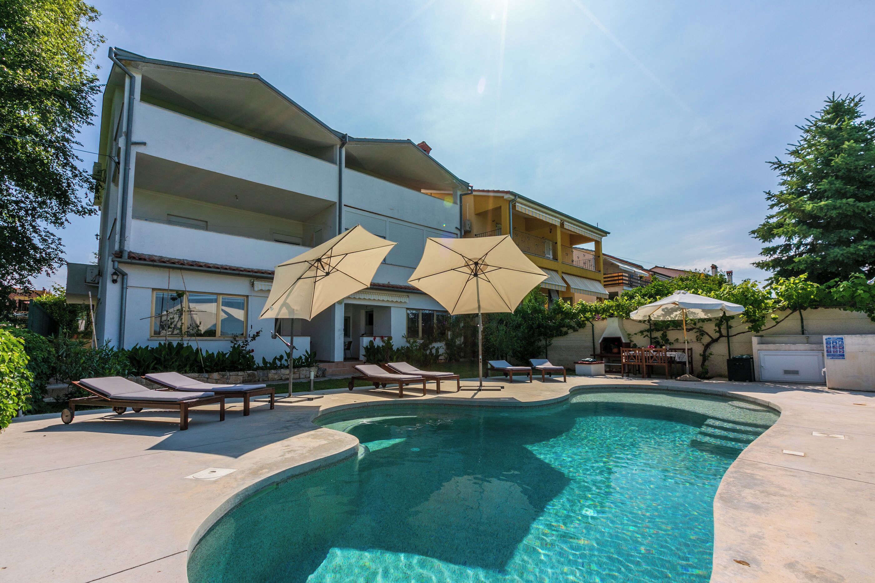 A swimming pool with lounge chairs and umbrellas, surrounded by a building with balconies and outdoor seating.