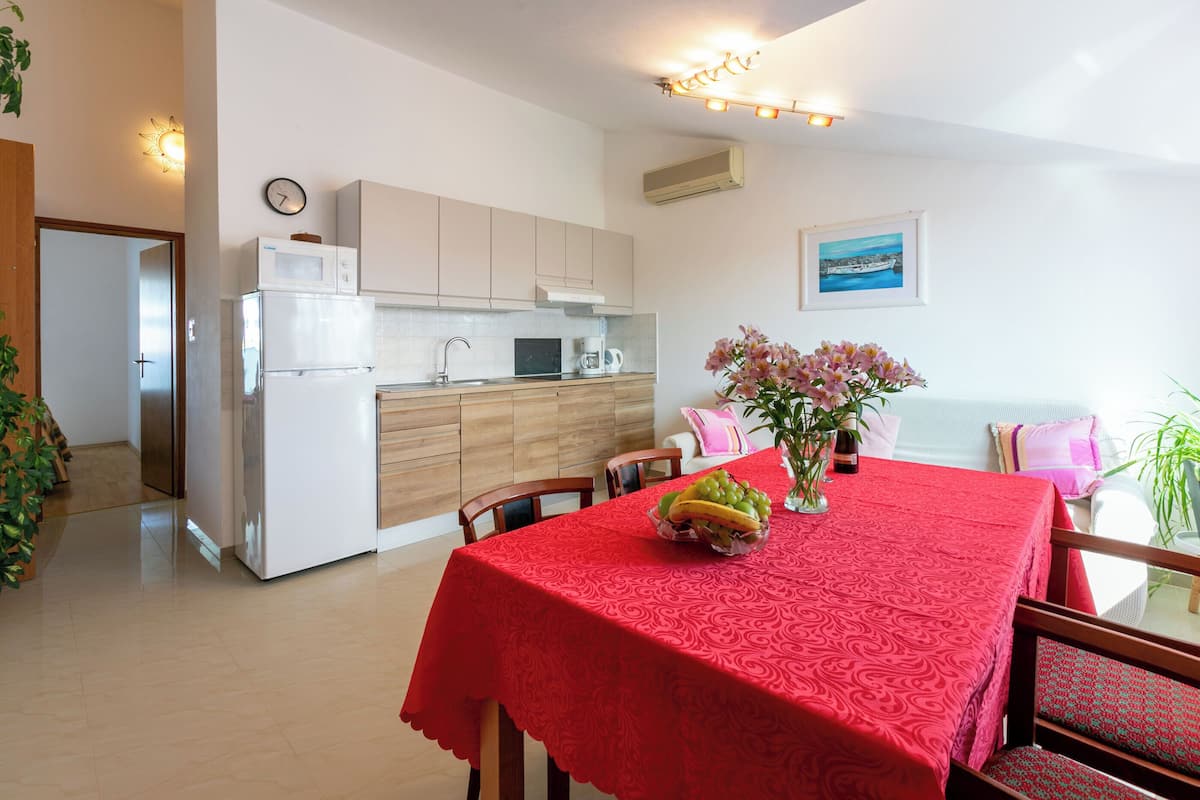A modern kitchen with white appliances, a dining area with a red tablecloth, and a view of the sea through a window.