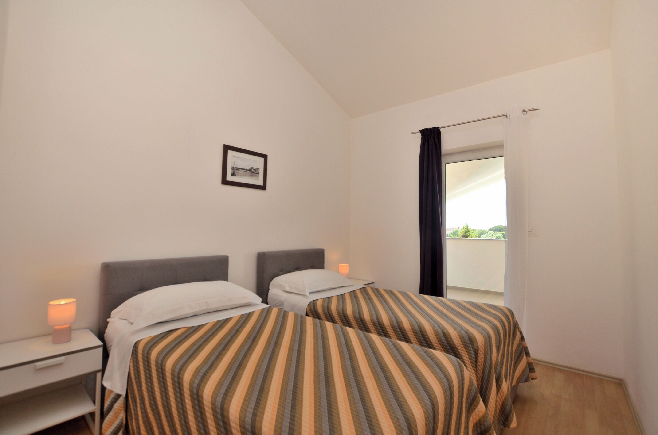 A bedroom with two beds, a nightstand with a lamp, a framed picture on the wall, and a window with a curtain.