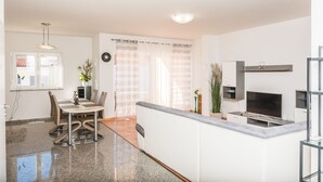 A modern kitchen with a built-in stove, a dining area with a table and chairs, and a large window with sheer curtains. - Apartments Marcel (Novi Vinodolski)
