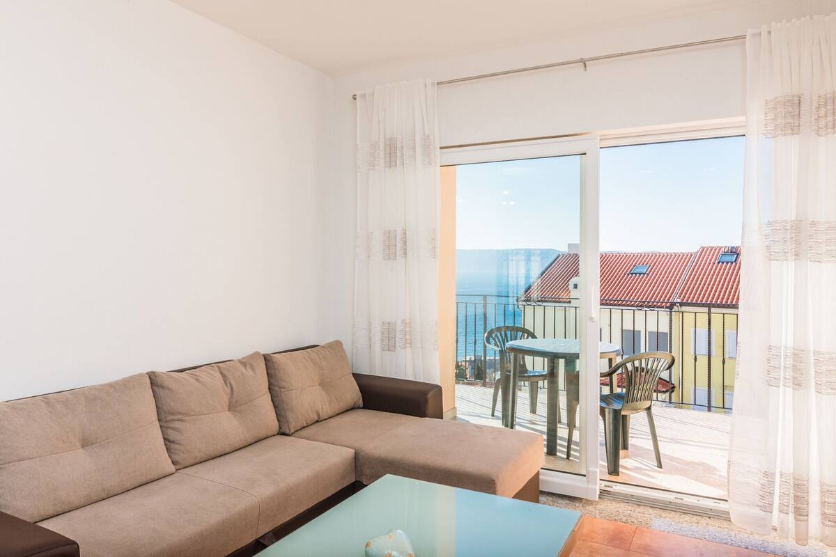 A modern living room with a sofa and a glass coffee table, a balcony with a table and chairs, and a view of buildings and a clear sky.