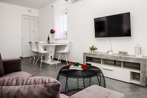 Una sala de estar moderna con un televisor de pantalla plana, un comedor con sillas blancas y una pequeña mesa redonda con un libro y una flor.