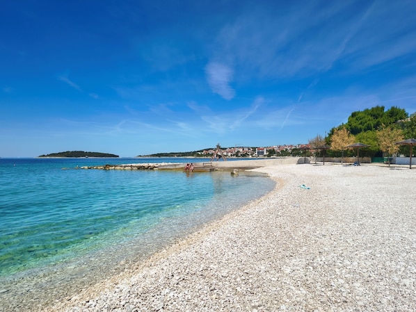 A pebble beach with clear water, a wooden swing, and a distant town.