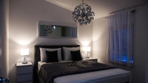 A modern bedroom with a large bed, two bedside tables with lamps, a unique chandelier, a mirror above the bed, and a window with blinds. - Apartments Zdeslav (Rogoznica)