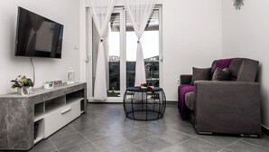 A modern living room with a flat-screen TV, a grey sofa, a round black side table, and a sliding glass door with sheer curtains. - Apartments Zdeslav (Rogoznica)