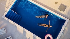 Vista aérea de una piscina rectangular con dos personas flotando, un flotador con forma de rosquilla y sillas reclinables blancas alrededor de la piscina.
