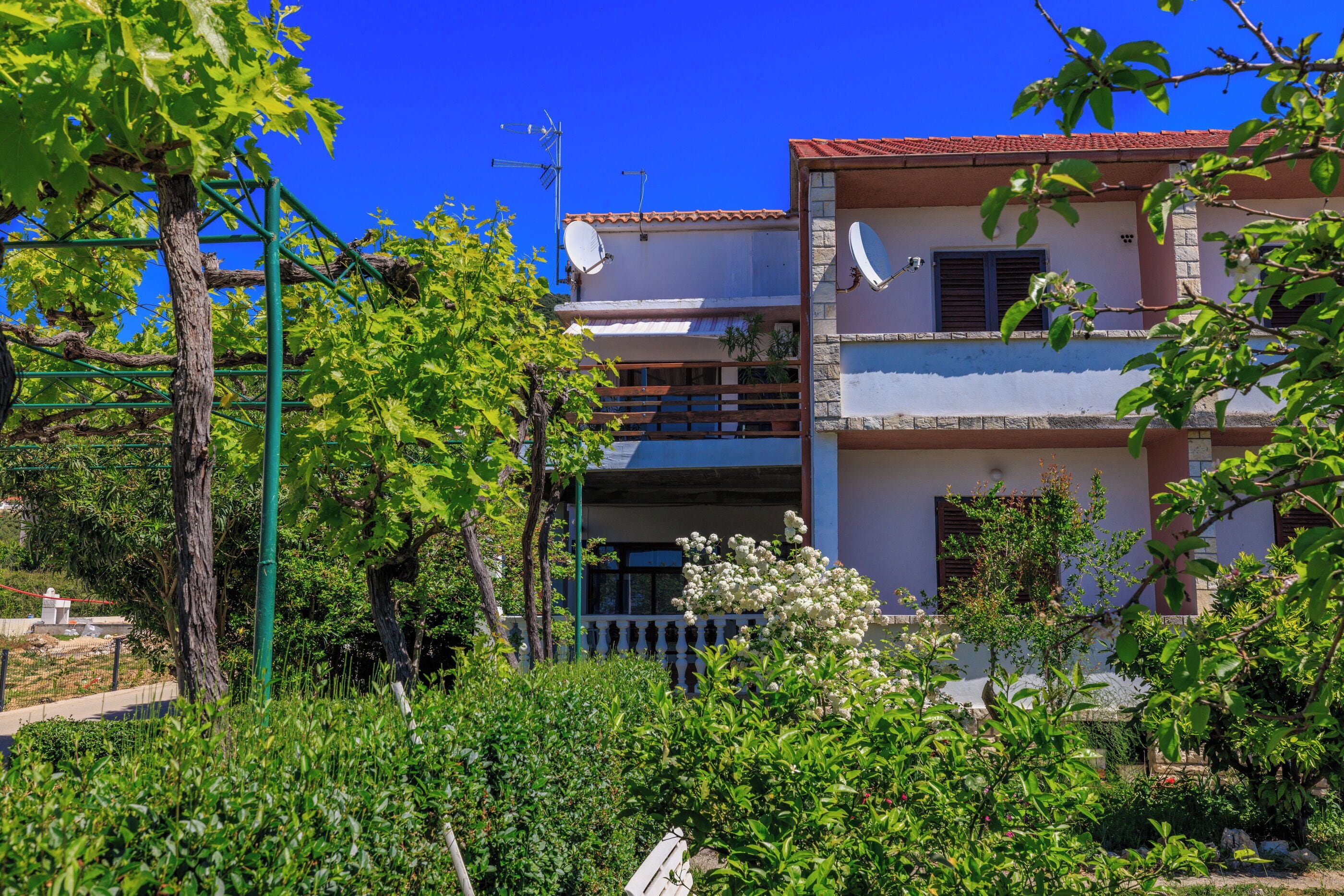 Edificio de dos pisos con antenas parabólicas, un balcón y un jardín con diversas plantas.