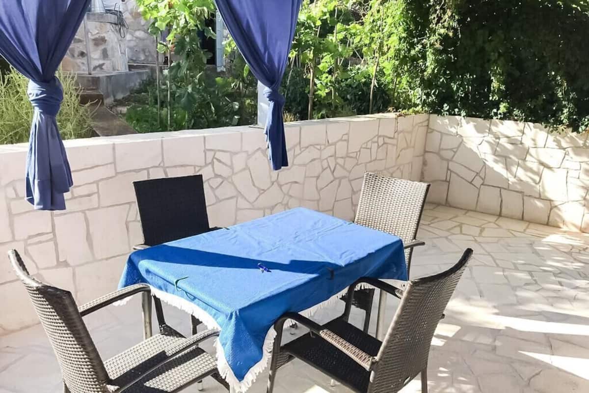 Un patio con una mesa y sillas, cubierto con un mantel azul, frente a una pared de piedra con follaje al fondo.