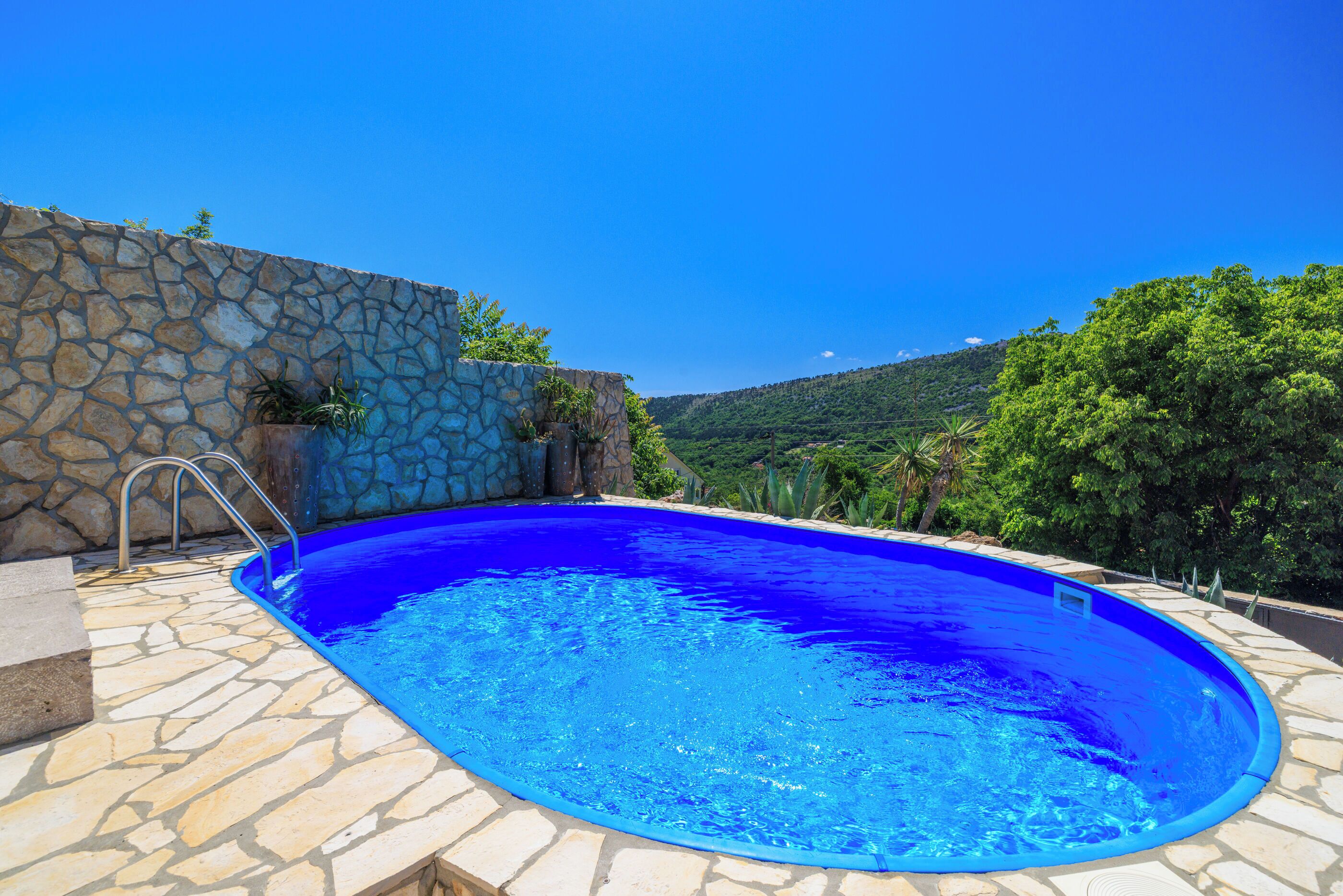 A swimming pool with stone walls and steps leading into it, surrounded by greenery and a clear sky.
