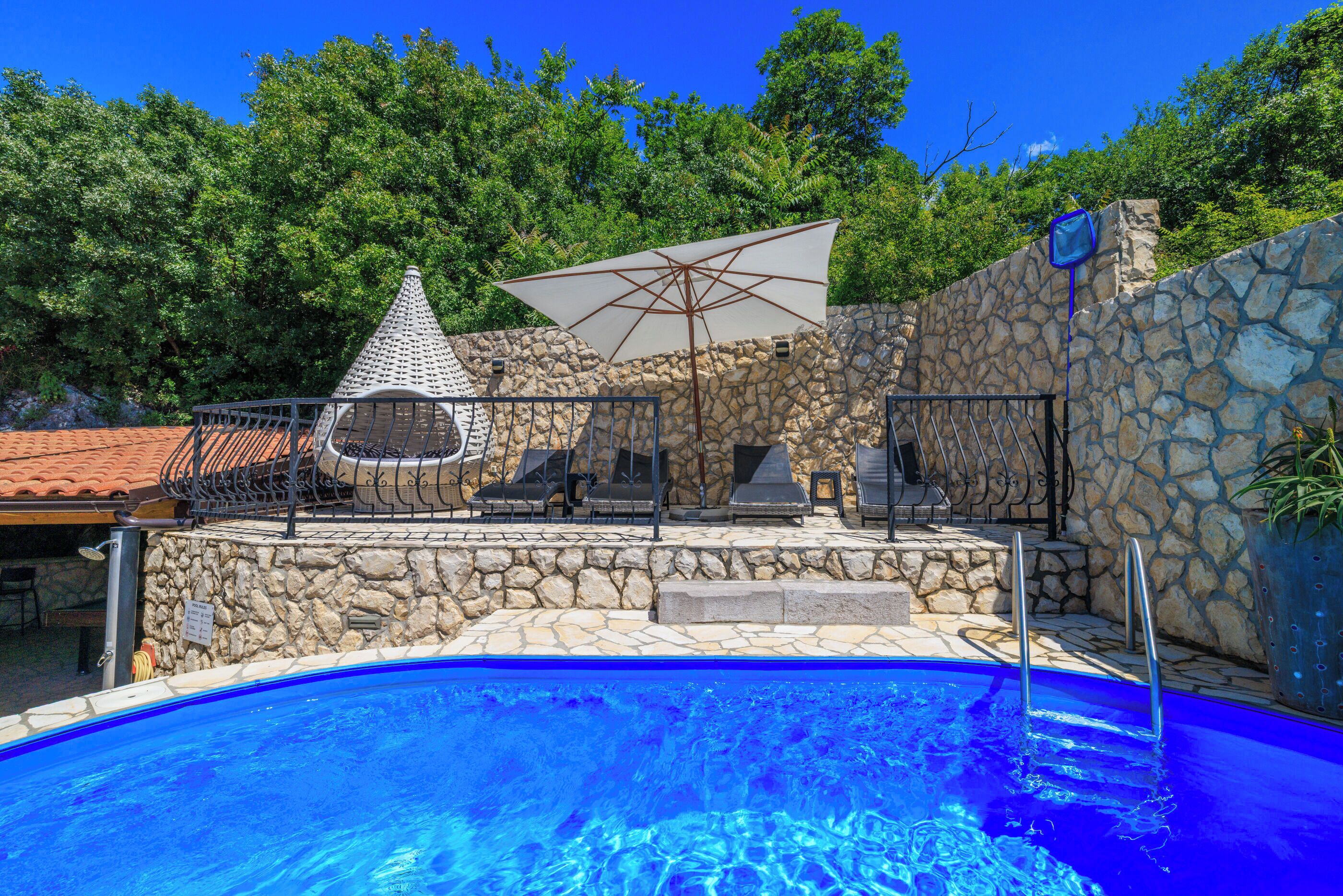 A swimming pool with a stone wall and a covered seating area with lounge chairs and an umbrella.