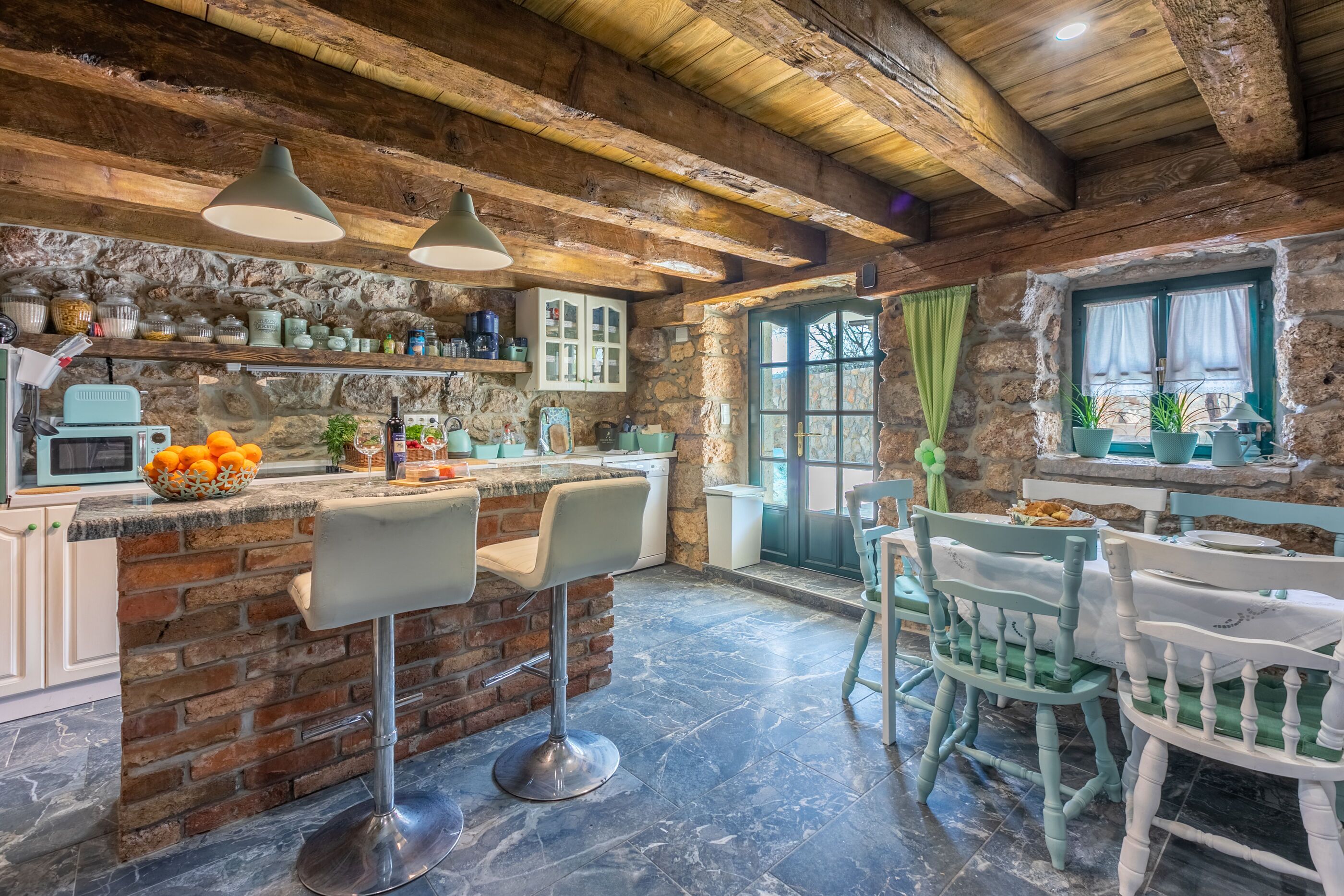 A rustic kitchen with exposed wooden beams, a stone wall, and a bar area with stools.