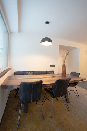 Un espace repas moderne avec une table en bois, des chaises noires, un vase contenant des fleurs séchées et un luminaire suspendu.