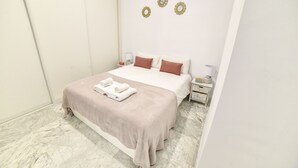 Een netjes opgemaakt bed met een beige sprei en twee kussens, een nachtkastje met een lamp en een wandklok.