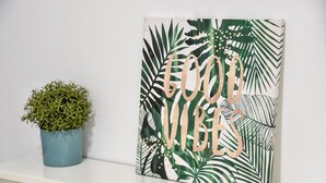 Een muur met een kunstwerk in een lijst, met tropische bladeren en de tekst 'GOOD VIBES'.