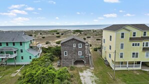 A coastal residential area with three houses, one with a balcony and a porch, another with a small porch, and a third with a large porch and a staircase leading to the entrance. - OCEANFRONT- Hatteras Island/Salvo: 2 Primaries, Hot Tub, Large Open Living Space (Salvo)