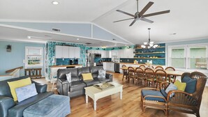 A spacious living room with a grey sofa, a white coffee table, and wicker chairs. A kitchen with white cabinets and a dining area with wooden tables and chairs is visible in the background. - OCEANFRONT- Hatteras Island/Salvo: 2 Primaries, Hot Tub, Large Open Living Space (Salvo)