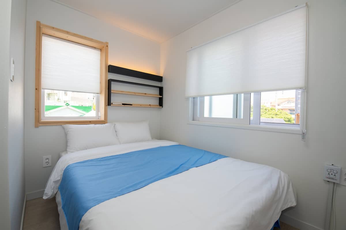A bedroom with a bed, a window with blinds, a shelf, and a power outlet.