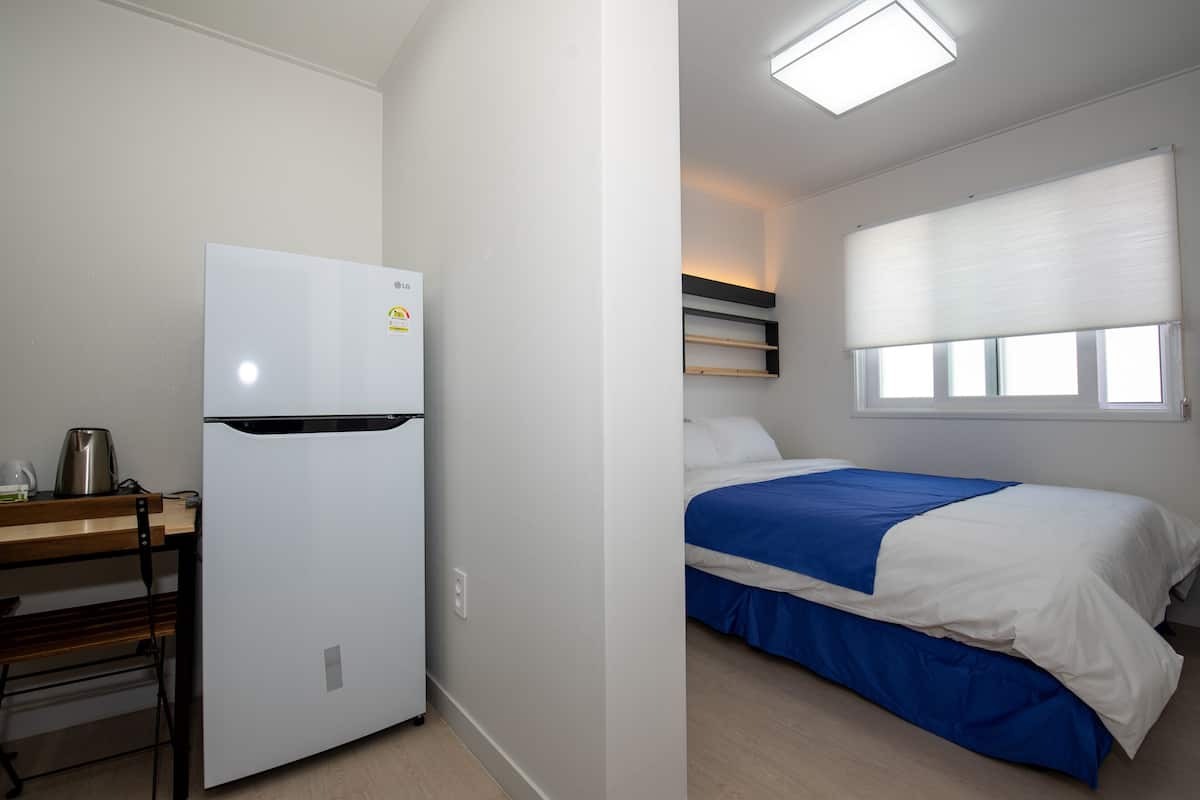 A compact room with a kitchenette area including a white refrigerator, a wooden table, and a kettle. There is a bed with blue bedding and a window with blinds.