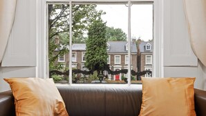 A room with a window view of a residential area, a brown leather sofa with two cushions, and a black metal gate. - Refined, Light 1 Bed Apt w Balcony in Brixton (London)