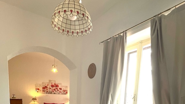 Amplia sala de estar con piso de madera en patrón chevron, un sofá blanco, un sillón rojo y un banco blanco. Un ventanal con cortinas grises deja entrar luz natural.