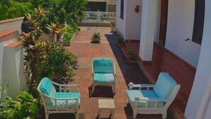 Un patio con paredes blancas, suelo de baldosas y algunas macetas. Hay dos sillas blancas y una pequeña mesa.