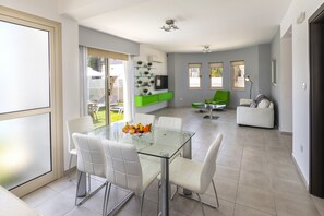 A modern living room with a glass dining table, white chairs, a green sofa, and a TV mounted on the wall.