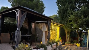 Un espace extérieur aménagé en patio, doté d’un coin salon couvert, de jardinières en pierre ornées de plantes et d’un mur décoratif illuminé.