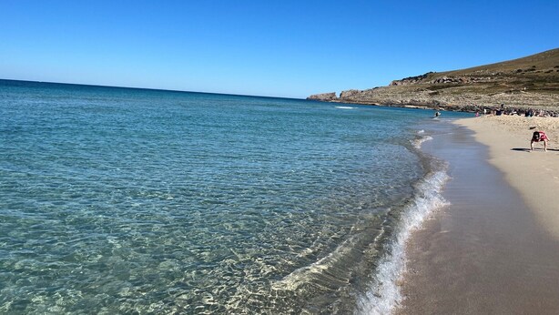 Une plage à l’eau claire, avec un rivage sablonneux et des falaises rocheuses en arrière-plan.