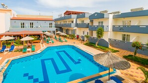 A swimming pool with a geometric design, surrounded by lounge chairs and umbrellas, and adjacent to multi-story buildings with balconies. - Anthoula Village (Hersonissos)