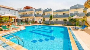 Una piscina con suelo azul, rodeada de sillas de descanso y sombrillas, con edificios de varios pisos y balcones aledaños.