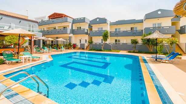 A swimming pool with a blue deck, surrounded by lounge chairs and umbrellas, and adjacent multi-story buildings with balconies. - Anthoula Village (Hersonissos)