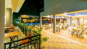 Un área de comedor al aire libre con mesas y sillas, una piscina al fondo y un edificio de varios niveles.